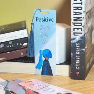 May include: A set of four bookmarks with colorful designs and tassels. The bookmarks feature illustrations of women and inspirational quotes. The quotes include "Positive Thoughts Vibes Life" and "Open a Book to find the Pearl Inside".