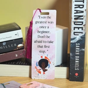 May include: Pink and white bookmark with a quote about being a beginner. The quote reads "Even the greatest was once a beginner. Don't be afraid to take that first step." The bookmark features a black woman with a pink and blue fish illustration.
