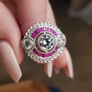 Puede incluir: Un anillo de plata con una piedra central redonda grande rodeada de piedras más pequeñas y una banda de piedras rosas.