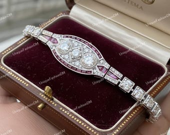 Op de afbeelding: Een Art Deco-armband met een centraal ovaal ontwerp met drie grote, ronde, heldere stenen en kleinere vierkante en rechthoekige stenen. De armband is bezet met kleinere heldere stenen en rode stenen en wordt gepresenteerd in een bruine sieradendoos.
