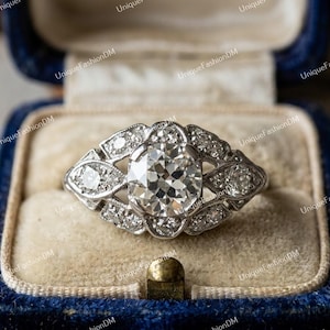 May include: A silver-toned ring featuring a large, round, faceted center stone surrounded by a decorative, openwork metal setting encrusted with smaller, clear, round-cut stones arranged in a symmetrical, elongated, marquise-like pattern.