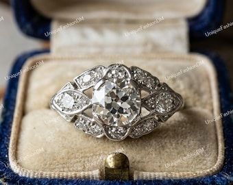 May include: A silver-toned ring featuring a large, round, faceted center stone surrounded by a decorative, openwork metal setting encrusted with smaller, clear, round-cut stones arranged in a symmetrical, elongated, marquise-like pattern.