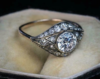 May include: A vintage-style ring with a large, round, clear diamond as the focal point. The ring features a gold band and is encrusted with smaller diamonds along the sides, set in an ornate, Art Deco-inspired design. The ring is displayed in a velvet ring box.