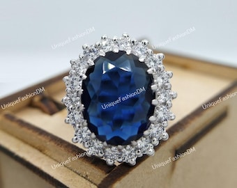 Op de afbeelding: Een zilveren ring met een grote, ovale, diepblauwe edelsteen omringd door kleinere, heldere stenen. De ring wordt gepresenteerd op een houten oppervlak, wat het ontwerp en de schittering van het sieraad benadrukt.