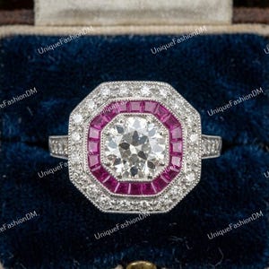 May include: An octagonal ring featuring a central round brilliant-cut diamond surrounded by a halo of square-cut pink rubies and an outer border of small round diamonds set in a silver-toned metal band with intricate milgrain detailing.