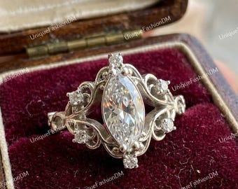May include: A marquise-cut diamond ring with a filigree design. The ring features a large, central diamond and smaller accent diamonds. The ring is set in a silver-colored metal and is displayed in a vintage jewelry box.