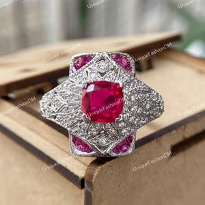 Peut inclure: Une bague en argent avec une grande pierre rouge de forme carrée entourée de petites pierres claires. La bague a un design délicat et complexe.