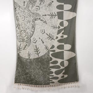 Könnte beinhalten: Ein grau-weißes Strandtuch mit Schildkrötenmuster. Das Tuch zeigt auf einer Seite eine große Schildkrötenillustration mit dekorativen Mustern und Formen. Die andere Seite hat ein geometrisches Muster. Der untere Rand hat eine Fransen.