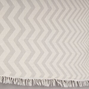 May include: A light gray and white chevron patterned textile with a fringed edge. The zigzag design runs vertically across the fabric. The textile appears to be made of a soft, woven material.