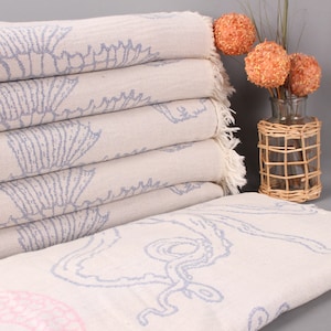 Peut inclure: Une pile de serviettes de plage beige clair avec un motif de coquillage bleu. Les serviettes ont des bords frangés et sont pliées avec soin. Un vase décoratif avec des accents floraux orange est à droite.
