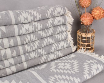 May include: A stack of gray and white patterned towels with fringed edges. The towels feature a geometric design and are neatly arranged. A decorative vase with orange accents is in the background.