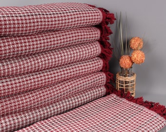 May include: A stack of burgundy and white patterned towels with fringed edges. The towels are neatly folded, showcasing a geometric design. A decorative vase with orange spheres and tall grass adds a touch of color.