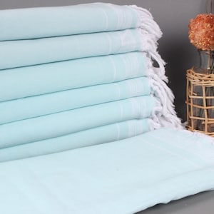 Puede incluir: Una pila de toallas de playa azul claro con flecos blancos. Las toallas están dobladas cuidadosamente, con rayas blancas cerca del borde superior. Un jarrón de cristal transparente con flores decorativas naranjas y una cesta de mimbre están en el fondo.