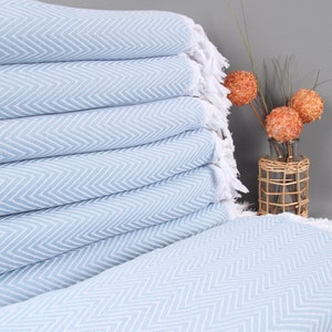 May include: A stack of light blue and white herringbone patterned towels with white fringe. The towels are neatly folded, showcasing the intricate weave. Decorative orange spheres and a vase are in the background.