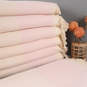 May include: A stack of light pink and cream-colored diamond-patterned towels with fringed edges. The towels are neatly folded, showcasing a textured weave. A decorative vase with orange spheres and a wicker basket adds a touch of decor.