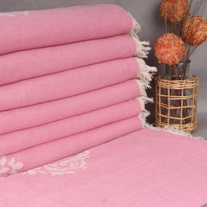 May include: A stack of pink towels with a white floral design on the edge. The towels have a fringed edge. A decorative vase with a woven basket around it and decorative balls is in the background.