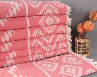 Puede incluir: Una pila de toallas turcas de color coral con patrones geométricos blancos y bordes con flecos. Las toallas están cuidadosamente dobladas, mostrando el intrincado diseño. Un jarrón decorativo con esferas naranjas también es visible.