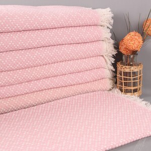 May include: A stack of pink and white patterned towels with fringed edges. The towels are folded neatly, with a matching towel spread out in front. A decorative vase with orange floral accents is in the background.