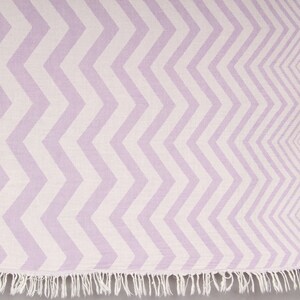 Puede incluir: Una toalla con estampado de chevron lavanda y blanco con un borde con flecos. El diseño en zigzag cubre toda la superficie de la toalla, creando una estética visualmente atractiva y moderna. La toalla está hecha de un material suave y tejido.