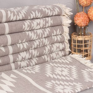 May include: A stack of folded, taupe-colored towels with a geometric white pattern. The towels have fringed edges and are neatly arranged. Decorative orange spheres in a glass vase are in the background.