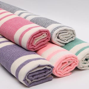 May include: Rolled-up striped towels in shades of pink, purple, gray, and green. Each towel features a white stripe design. The towels are made of a soft, woven material, perfect for the bathroom or beach.