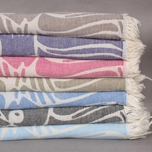 May include: A stack of colorful, patterned towels in shades of brown, purple, fuchsia, khaki green, light blue, black, and dark baby blue. Each towel features a white wave-like design and a fringed edge. The towels are neatly folded.