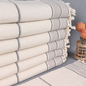 May include: A stack of cream-colored Turkish towels with gray and black striped accents. The towels have fringed edges and are neatly folded. The image also includes a decorative vase with orange floral accents.