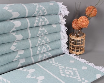 May include: A stack of six light teal-colored towels with white geometric patterns and fringed edges. The towels are folded neatly, showcasing the design. A decorative vase with orange decorative balls is in the background.