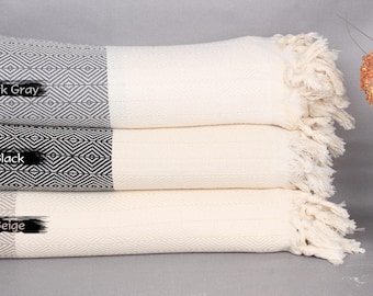 May include: Three Turkish towels stacked on top of each other. The top towel is beige, the middle towel is black with a diamond pattern, and the bottom towel is dark grey with a diamond pattern. Each towel has a fringe on the edges.