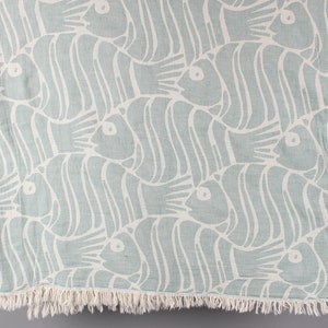 May include: A light blue and white patterned beach towel featuring a repeating fish design. The fish are stylized with curved lines and eye details. The towel has a fringed edge.