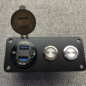 May include: Black panel with a silver USB charging port with two ports and the text "DC QC 3.0" on either side. There are two silver push buttons to the right of the charging port.