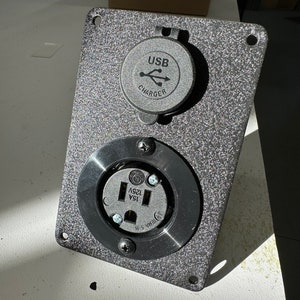 May include: A black metal electrical outlet plate with a standard 120V outlet and a USB charger. The plate has a textured surface and is labelled "15A 125V" and "USB CHARGER".