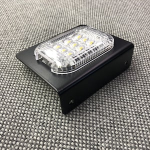 May include: A black metal bracket with a clear plastic LED light fixture. The light fixture has nine white LEDs.