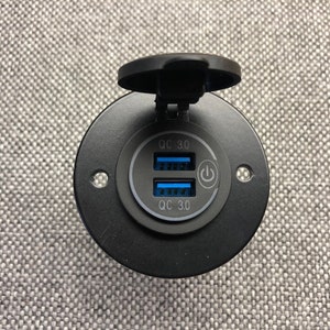 May include: A black USB charging port with two QC 3.0 ports and a power button symbol. The port has a protective cover and is designed for flush mounting. The background is a textured gray surface.