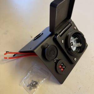 May include: A black electrical power panel featuring a large circular outlet with a hinged cover, a smaller circular port with a rubber cap, and a dual-terminal connector. Two red wires extend from the side, and a small plastic bag of screws is included.