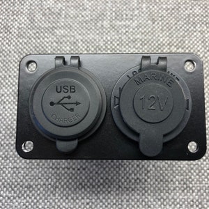 May include: Black rectangular panel with two electrical outlets. One outlet is labeled "USB CHARGER" and the other is labeled "MARINE 12V".