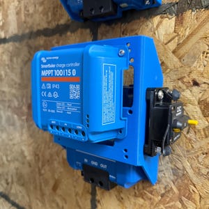 May include: A blue SmartSolar charge controller MPPT 100/15 with text and a QR code. The controller is mounted on a wooden surface. The controller has multiple ports and indicator lights. A black circuit breaker is attached to the side.