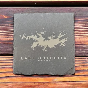 May include: A gray slate coaster with a white outline of Lake Ouachita, Arkansas. The text "LAKE OUACHITA" and "ARKANSAS" is printed in white below the lake outline.