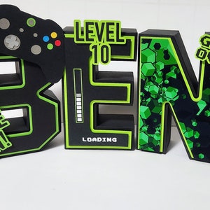 May include: Black and green foam letters spelling "BEN" with a green and black video game controller, a green and black "LEVEL 10" sign, a green and black "GAME ON!" sign, and a green and black "GAME OVER" sign. The letters have a green outline and a black background with a green pixelated pattern.
