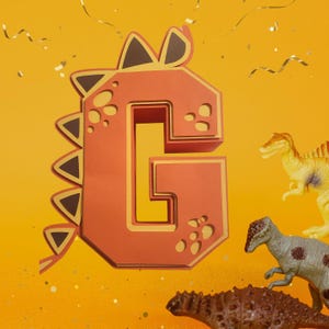Könnte beinhalten: Ein großer, dreidimensionaler, orangefarbener Papierbuchstabe G in Form eines Dinosauriers. Der Buchstabe ist mit braunen Stacheln und gelben Akzenten verziert. Der Buchstabe ist von Konfetti und Spielzeug-Dinosauriern umgeben.