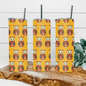 May include: Three stainless steel tumblers with a yellow background and a repeating pattern of red, brown, and blue owls. The tumblers are on a wooden surface.