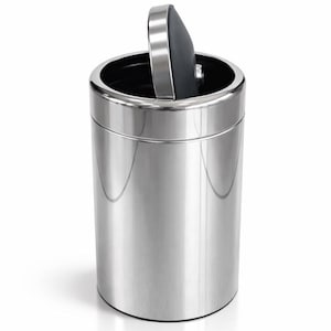 Puede incluir: Un cubo de basura cilíndrico con un acabado cromado pulido y una superficie metálica reflectante. El recipiente presenta una abertura circular con una tapa basculante negra en forma de semicírculo, parcialmente abierta, que revela el interior oscuro.