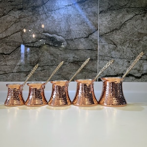 Könnte beinhalten: Fünf Kupfer-Kaffeekannen mit Henkeln, jede mit einer gehämmerten Textur. Die Kannen sind in einer Reihe von der größten bis zur kleinsten angeordnet.