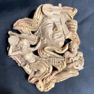 Op de afbeelding: Een beige keramische wandsculptuur die een gezicht afbeeldt dat omringd is door muziekinstrumenten, waaronder een viool, een trompet en een fluit. De sculptuur is gedetailleerd met ingewikkelde houtsnijwerk en een getextureerd oppervlak.