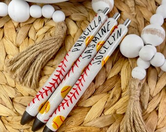 May include: Three baseball-themed pens with white glitter barrels, red stitching, and yellow baseball designs. Each pen is labeled with a name: Priscilla, Jasmine, and Jocelyn. The pens are set on a woven, light brown surface.