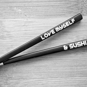 Puede incluir: Un par de palillos chinos negros con letras blancas. Uno dice "LOVE MYSELF" y el otro "& SUSHI". Están colocados sobre un fondo de madera. Un accesorio elegante y divertido para disfrutar del sushi.