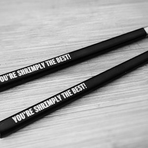Puede incluir: Dos palillos chinos negros con puntas plateadas, cada uno con el texto blanco "YOU'RE SHRIMPPLY THE BEST!". Los palillos están dispuestos en diagonal sobre una superficie de madera clara, creando un diseño juguetón.