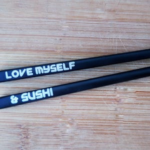 Puede incluir: Dos palillos negros con puntas plateadas sobre una superficie de madera. Uno tiene el texto "LOVE MYSELF" en blanco, y el otro "& SUSHI". Estos palillos son un accesorio divertido para disfrutar del sushi.