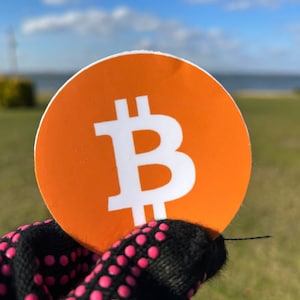 Op de afbeelding: Een close-up van een ronde oranje sticker met een wit Bitcoin-symbool erop. De sticker wordt vastgehouden door een hand die een zwarte handschoen met roze stippen draagt.
