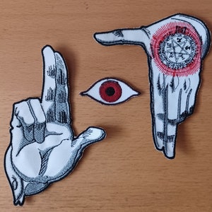 May include: Three embroidered patches featuring two white hands with black line shading and a central red and white eye. One hand displays a circular red and black sigil with a pentagram. The patches have black borders and white fabric bases.
